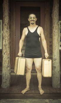 Goofy Gentleman Dressed in 1920’s Era Swimsuit Holding Suitcases on Porch of Cabin.