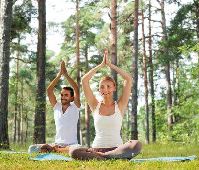 sport, fitness, yoga and people concept - smiling couple meditating and sitting on mats with raised hands over green woods background