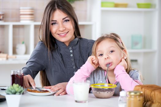 Mother and her daughter preparing breakfast. Mother smear jam on bread. Daughter fool around with cereal in milk. Looking at camera.