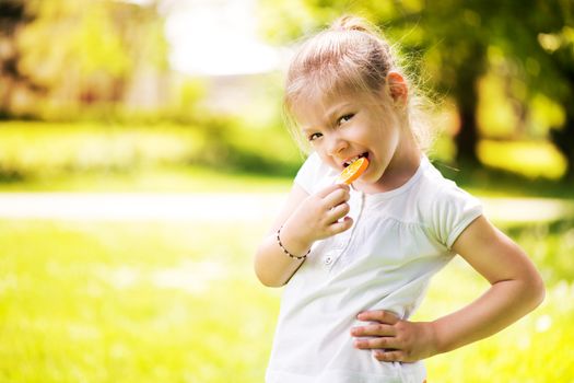 Cute Little Girl Standing on Green Grass in the Park and holding in hand orange Lollipop.