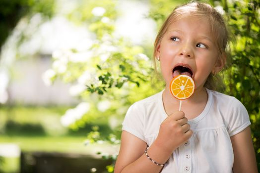 Portrait of Cute Little Girl Standing in the Park and holding in hand orange Lollipop.