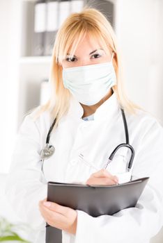 Portrait of a young woman doctor with stethoscope and surgical mask standing in her office and holding clipboard.