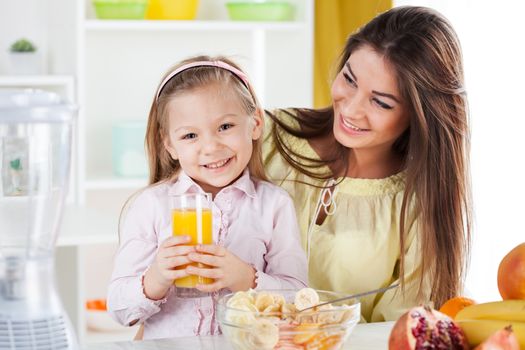 Mother and daughter in the kitchen sitting with table. Little girl holding orange juice. Fruit salad standing on table.