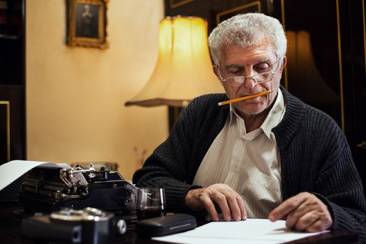 Retro Senior man writer with glasses and pencil in his mouth sitting at the desk and reading some text for writing on Obsolete Typewriter.