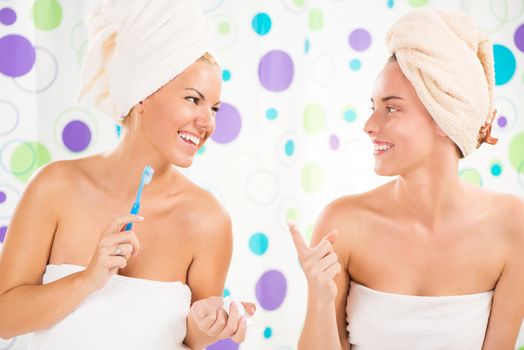 Two young cute woman preparing to start their day. One girl brushing teeth, the other girl applying cream on the face with finger.