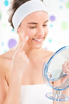 Young cute woman preparing to start her day. She is applying moisturizer cream on face. Looking at mirror.