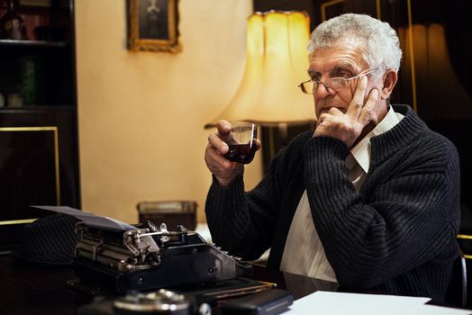 Retro Senior man writer with glasses sitting at the desk with glass of whiskey and thinking.