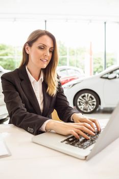 Smiling saleswoman typing on her laptop at new car showroom