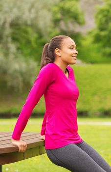 fitness, sport, training, park and lifestyle concept - smiling african american woman doing push-ups on bench outdoors