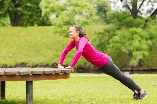 fitness, sport, training, park and lifestyle concept - smiling african american woman doing push-ups on bench outdoors