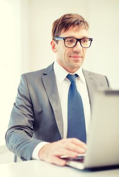 business, internet, technology and office concept - smiling businessman in eyeglasses with laptop computer in office