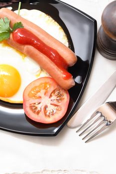 Black plate with fried eggs and sausages on white table cloth near fork and knife