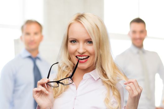 business, teamwork and people concept - smiling businesswoman, student or secretary with eyeglasses over office and group of colleagues background