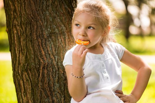 Cute Little Girl Standing in the Park leaning on a tree and holding in hand orange Lollipop.