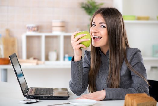 Businesswoman in the kitchen eating apple and reading mail on laptop before going to work.