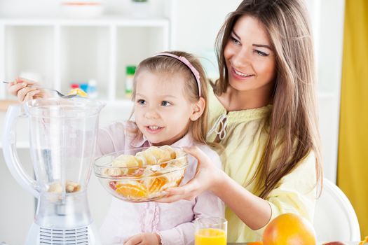 Beautiful young woman and cute little girl put the fruit in a blender in the kitchen.