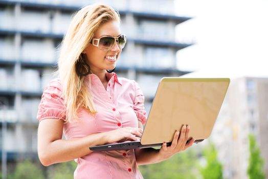 Beautiful young smiling business woman standing in front of building and using laptop outdoor.