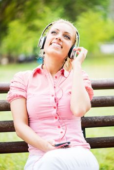Happy Young Woman sitting on the Park Bench and Listening Music from mobile phone.