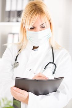 Portrait of a young woman doctor with stethoscope and surgical mask standing in her office and holding clipboard.