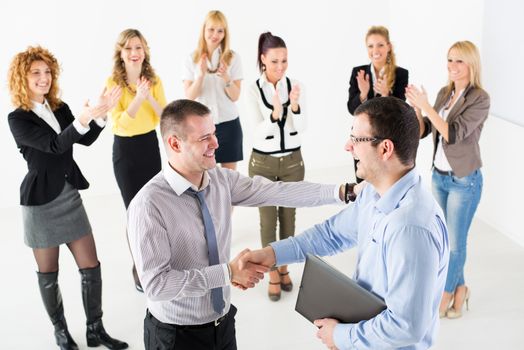 Two successful businessmen shaking hands at the meeting. Group of their colleague celebrating in the background.