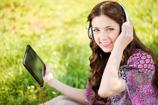 Happy Young Woman sitting on grass in the Beautiful Nature and Listening Music from digital tablet.