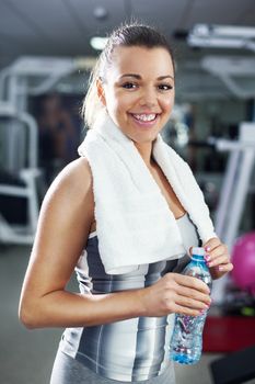 Cute Young girl standing and resting in modern gym and holding a bottle of water and towel. Looking At Camera.