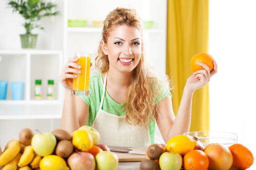 Beautiful young woman in the kitchen, holding orange and orange juice. Looking at camera.