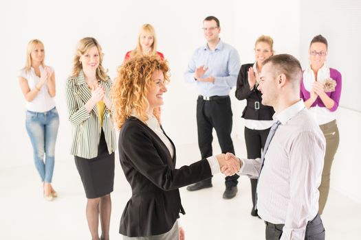 Two successful business people shaking hands at the meeting. Group their colleague celebrating in the background.