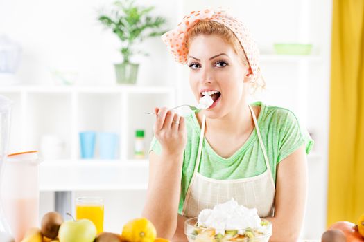 Beautiful young woman eat fruit salad in a kitchen. Looking at camera.