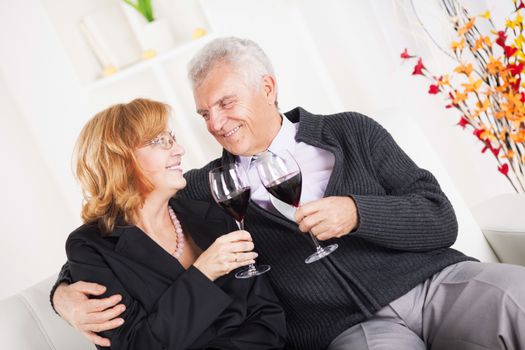 Happy senior couple sitting embraced at home, smiling and drinking red wine.