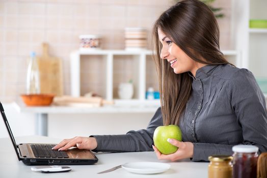 Businesswoman in the kitchen with apple reading mail on laptop before going to work.