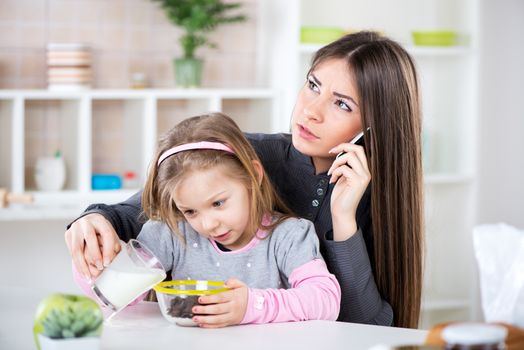 Overworked Business Woman and her little daughter in the morning. Overworked mother make phone calls before going to work. Preparing cereal with milk for her daughter and spilling milk next to bowl.