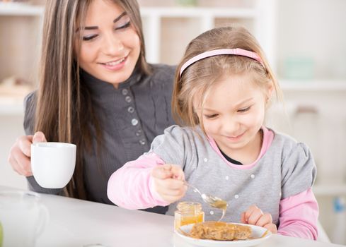 Mother and daughter breakfast in the kitchen. Cute little girl smear honey over the peanut butter on bread. Mother drinking coffe.