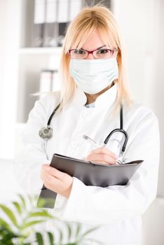 Portrait of a young woman doctor with stethoscope and surgical mask standing in her office and holding clipboard.