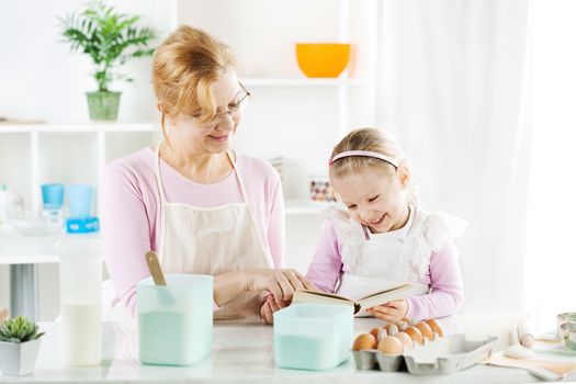 Beautiful happy grandmother and granddaughter reading cookery book in a kitchen. 