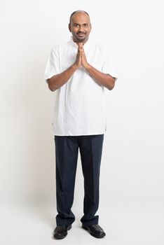 Portrait of full length mature casual business Indian man praying, standing on plain background with shadow.