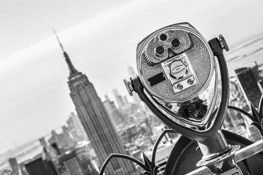 New York City, USA. Vintage tourist binoculars at Top of the Rock observation deck in front of Manhattan downtown skyline with Empire State Building and skyscrapers at sunset. Black and white.