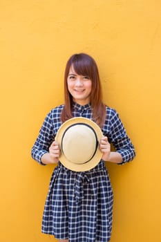 smiling woman wear in dress hand holding hat on yellow cement wall background