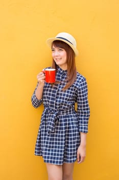 smiling woman wear in dress and hat holding red coffee cup on yellow cement wall background