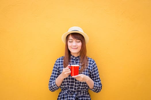 smiling woman wear in dress and hat holding red coffee cup on yellow cement wall background