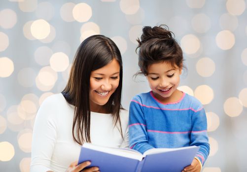 family, children, education and happy people concept - happy mother and little daughter reading book over holidays lights background