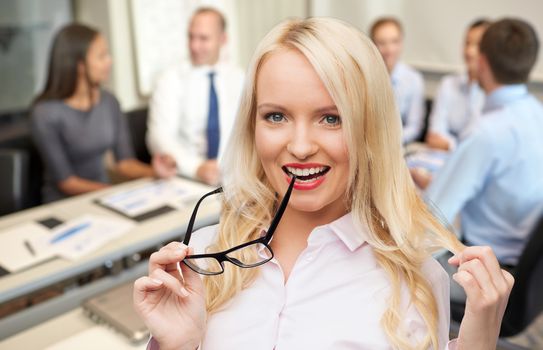 business, teamwork and people concept - smiling businesswoman, student or secretary with eyeglasses over office and group of colleagues background