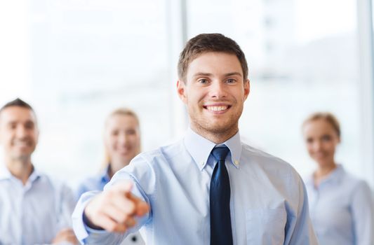 business, people, gesture and teamwork concept - smiling businessman pointing finger to you with group of businesspeople meeting in office