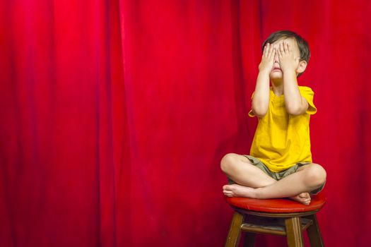 Mixed Race Boy Boy Covering His Eyes Sitting on Stool in Front of Red Curtain.