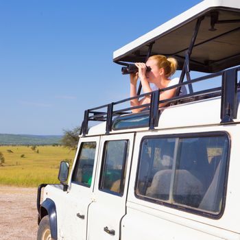 Young blond lady on safari standing in open roof jeep observing wild animals through binoculars.