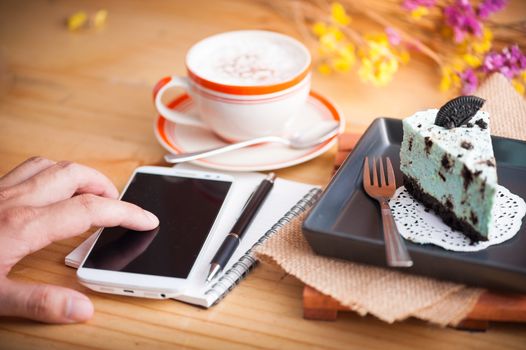 Male left hand finger touching on smartphone, peppermint with chocolate cookie cheese cake, notebook and pen, and hot cappuccino at cafe in afternoon time