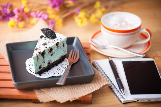 Peppermint with chocolate cookie cheese cake, notebook and pen, smartphone, and hot cappuccino at cafe in afternoon time