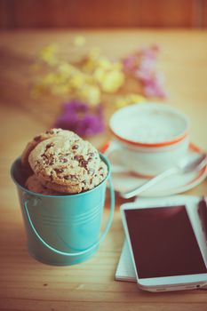 Chocolate chip cookies in small green bucket with smartphone, notebook and pen, and coffee cup at cafe in morning time with film filter effect