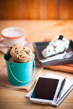 Chocolate chip cookies in small green bucket with smartphone, notebook and pen, and coffee cup at cafe in morning time