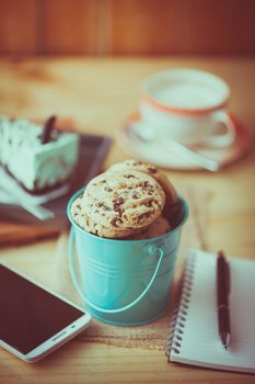 Chocolate chip cookies in small green bucket with smartphone, notebook and pen, and coffee cup at cafe in morning time with film filter effect
